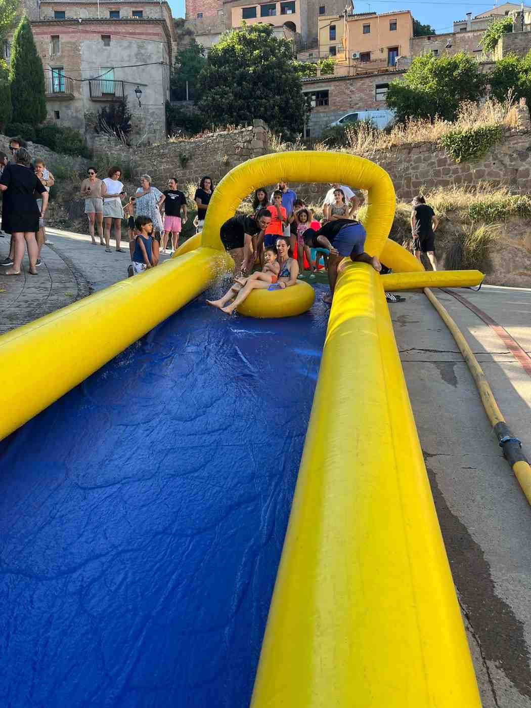 Gente deslizándose por un tobogán inflable amarillo hacia una piscina azul al aire libre.
