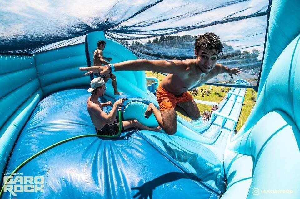 Niños deslizándose sobre un obstáculo acuático inflable azul con el océano y las montañas de fondo.