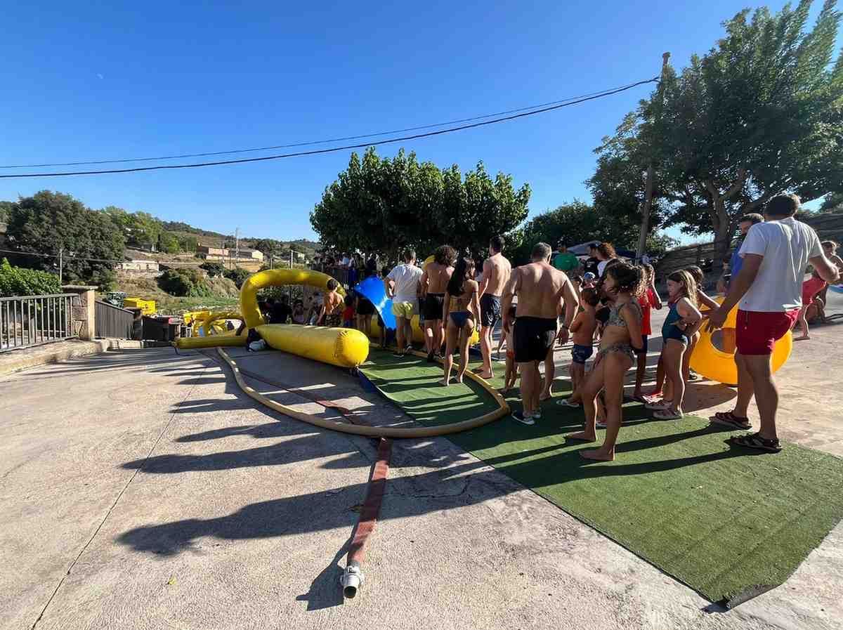 Gente esperando junto a un tobogán acuático amarillo en una soleada zona de parque al aire libre.