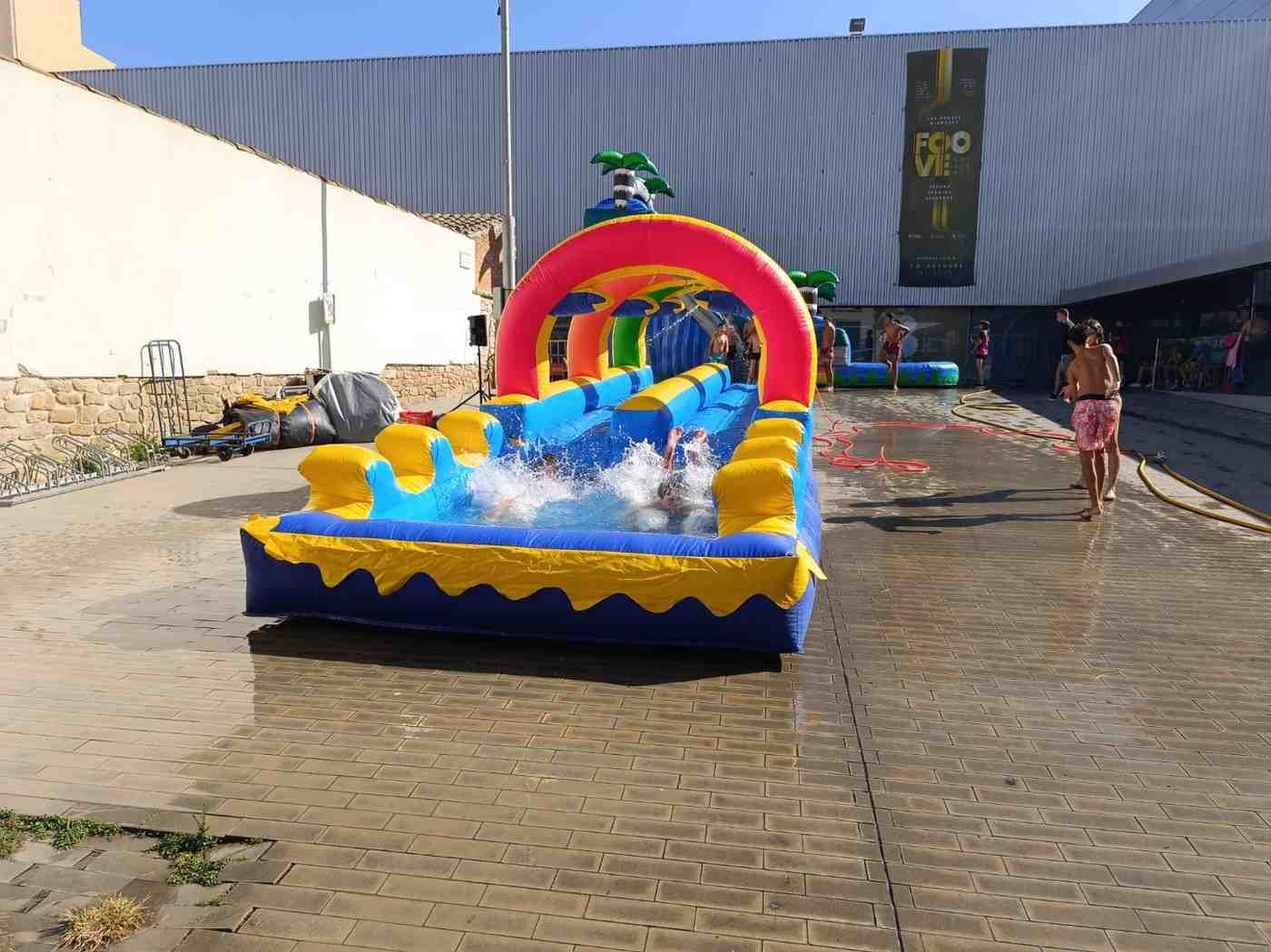 Tobogán acuático inflable de colores en un patio pavimentado iluminado por el sol con un niño cerca.