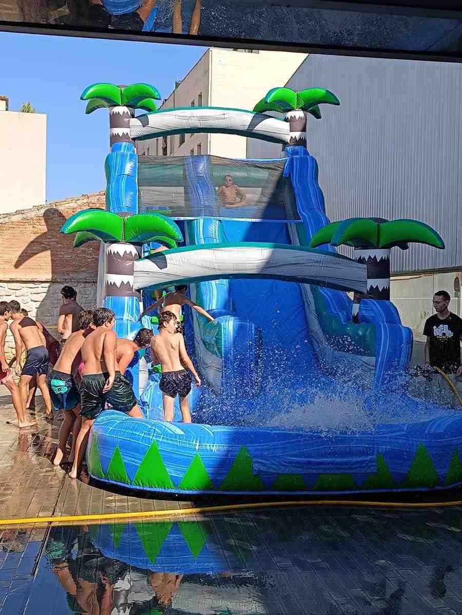 Niños jugando en un tobogán acuático azul con decoraciones de palmeras en una piscina al aire libre.