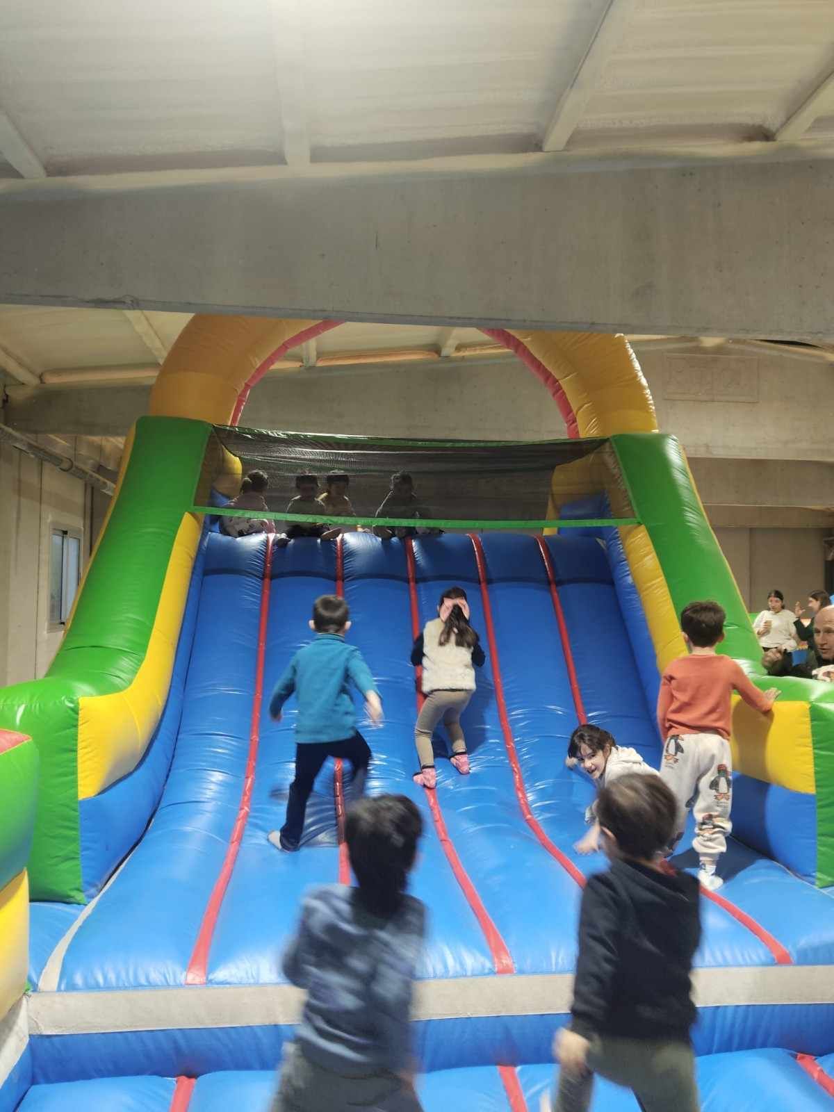Niños trepando por una colorida estructura de juegos inflable en el interior, con un tobogán azul.