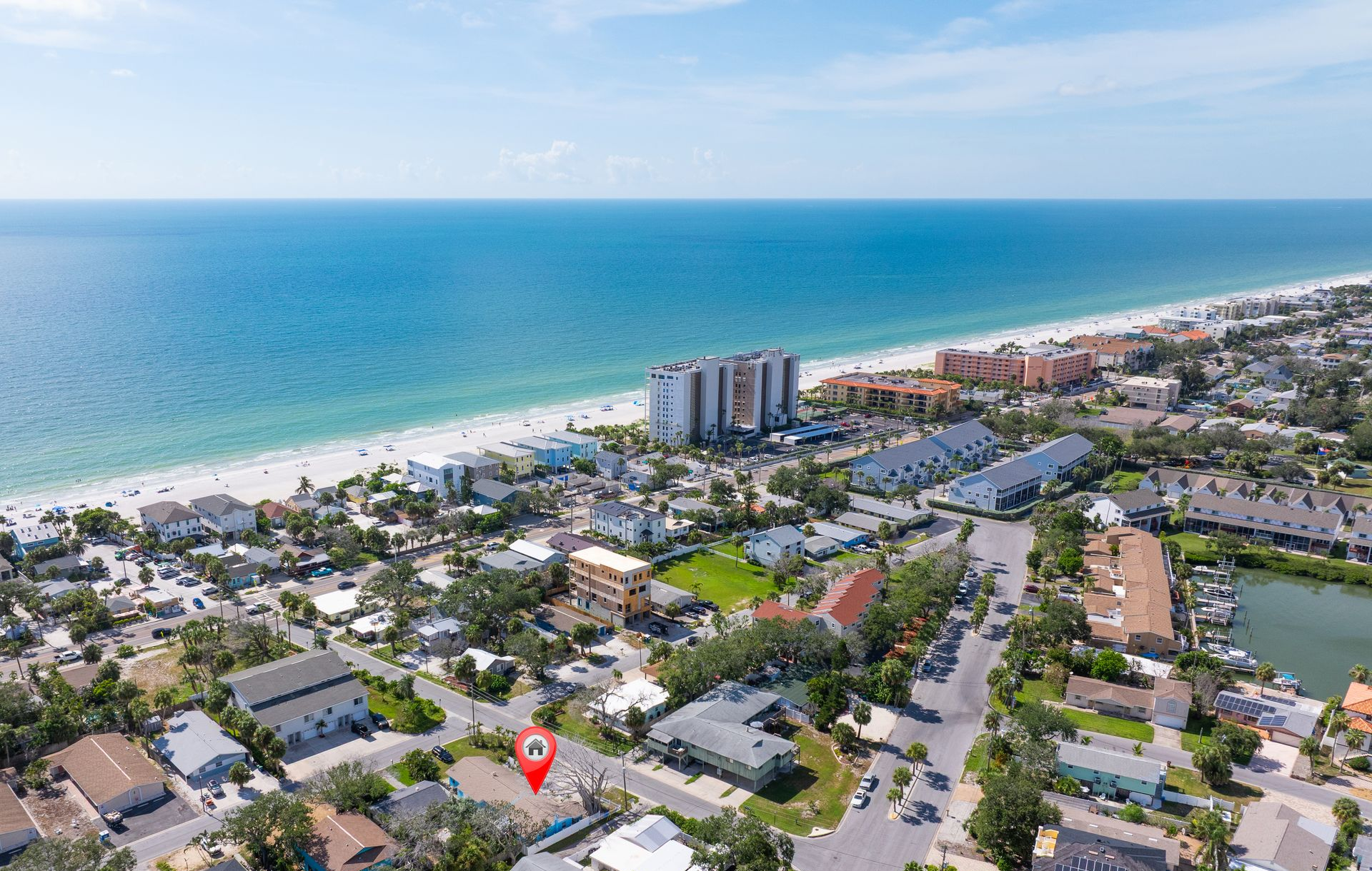 Aerial view of a coastal town with a beach, ocean, and buildings; a red location marker is in the foreground.