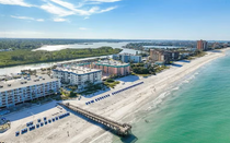 Aerial view of a sunny beach with white sand, turquoise water, and a pier beside coastal condos.