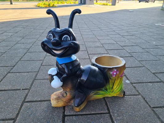Een zwart hondenbeeldje met gewei en blauwe halsband staat naast een bloempot op een geplaveide stoep.
