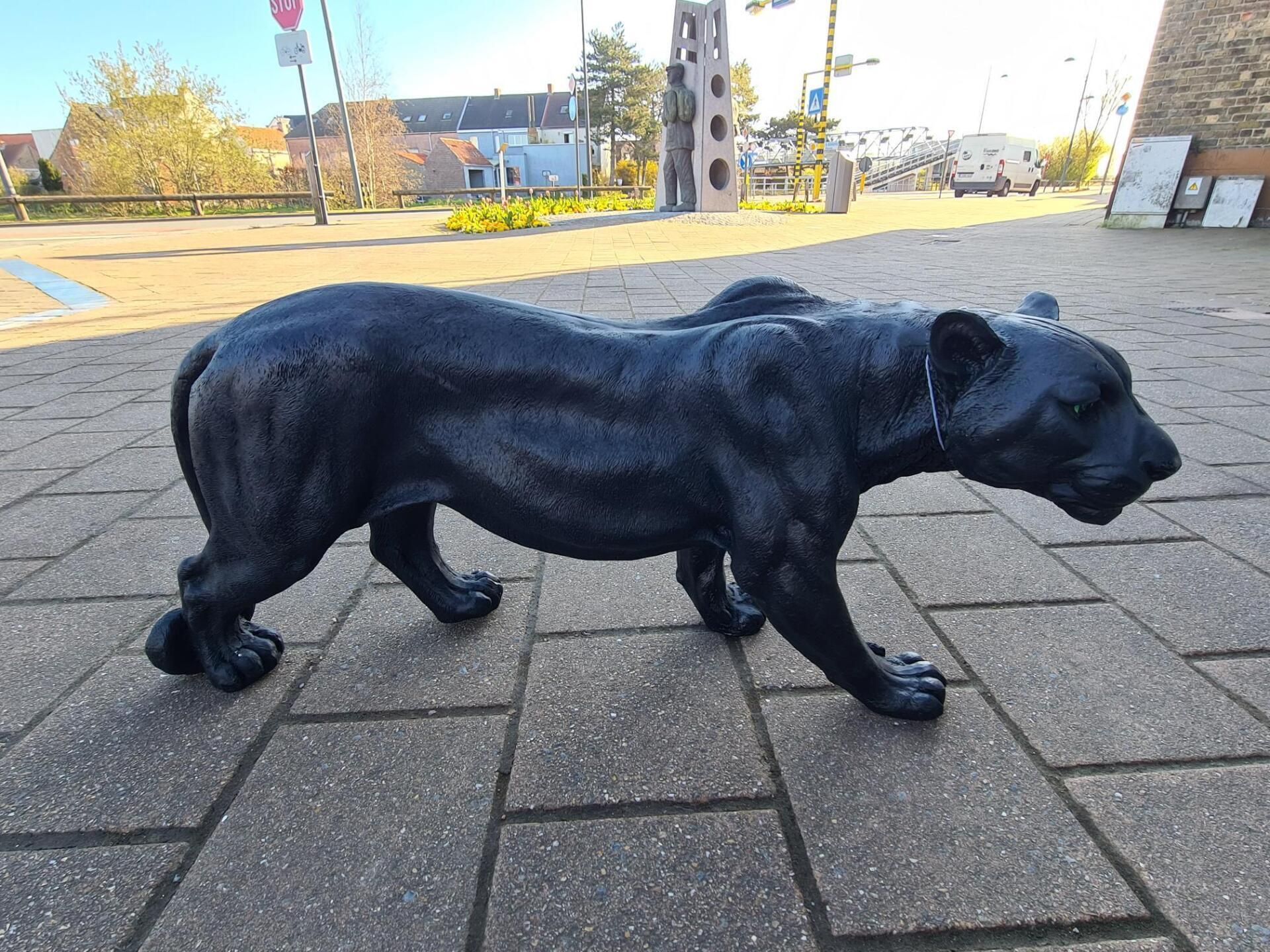 Standbeeld van een zwarte panter die over een geplaveid trottoir loopt, met gebouwen en bomen op de achtergrond.