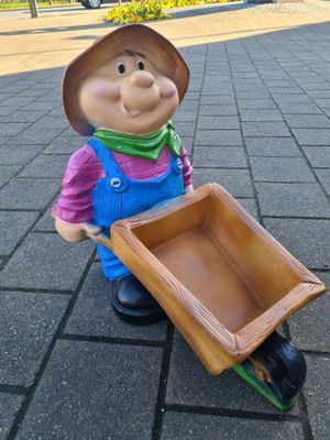 Een kleine tuinkabouter met een houten dienblad, een bruine hoed, een groene sjaal en een blauwe overall aan, staat buiten op de stoeptegels.