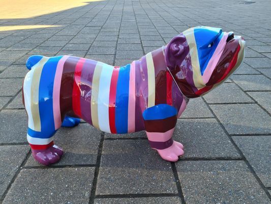 Kleurrijk gestreept bulldogbeeld op een betegelde stoep in de buitenlucht, geschilderd in blauw, roze, paars en rood.