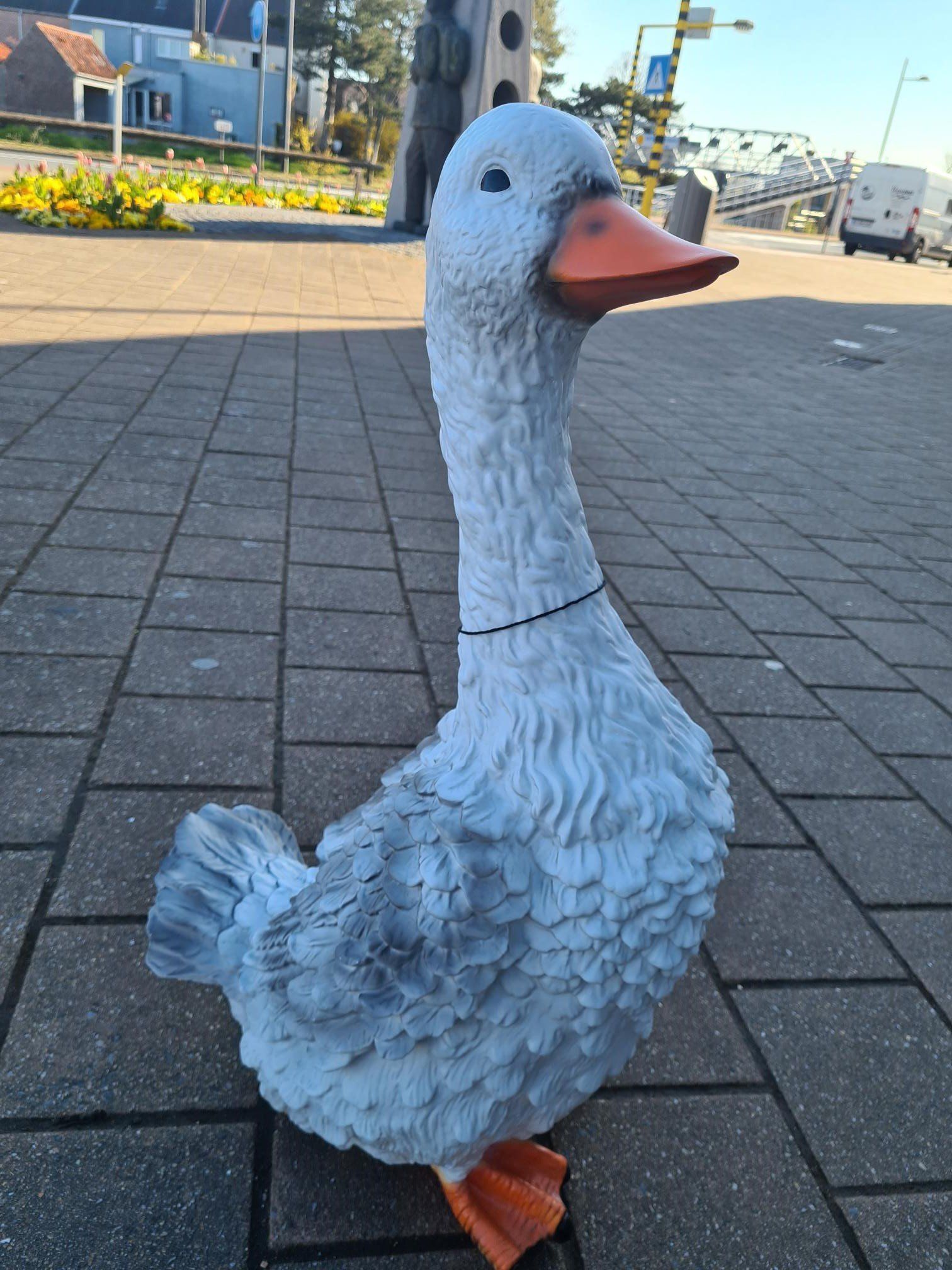 Een wit ganzenbeeld op een bakstenen pad bij bloemperken en gebouwen in de buitenlucht.