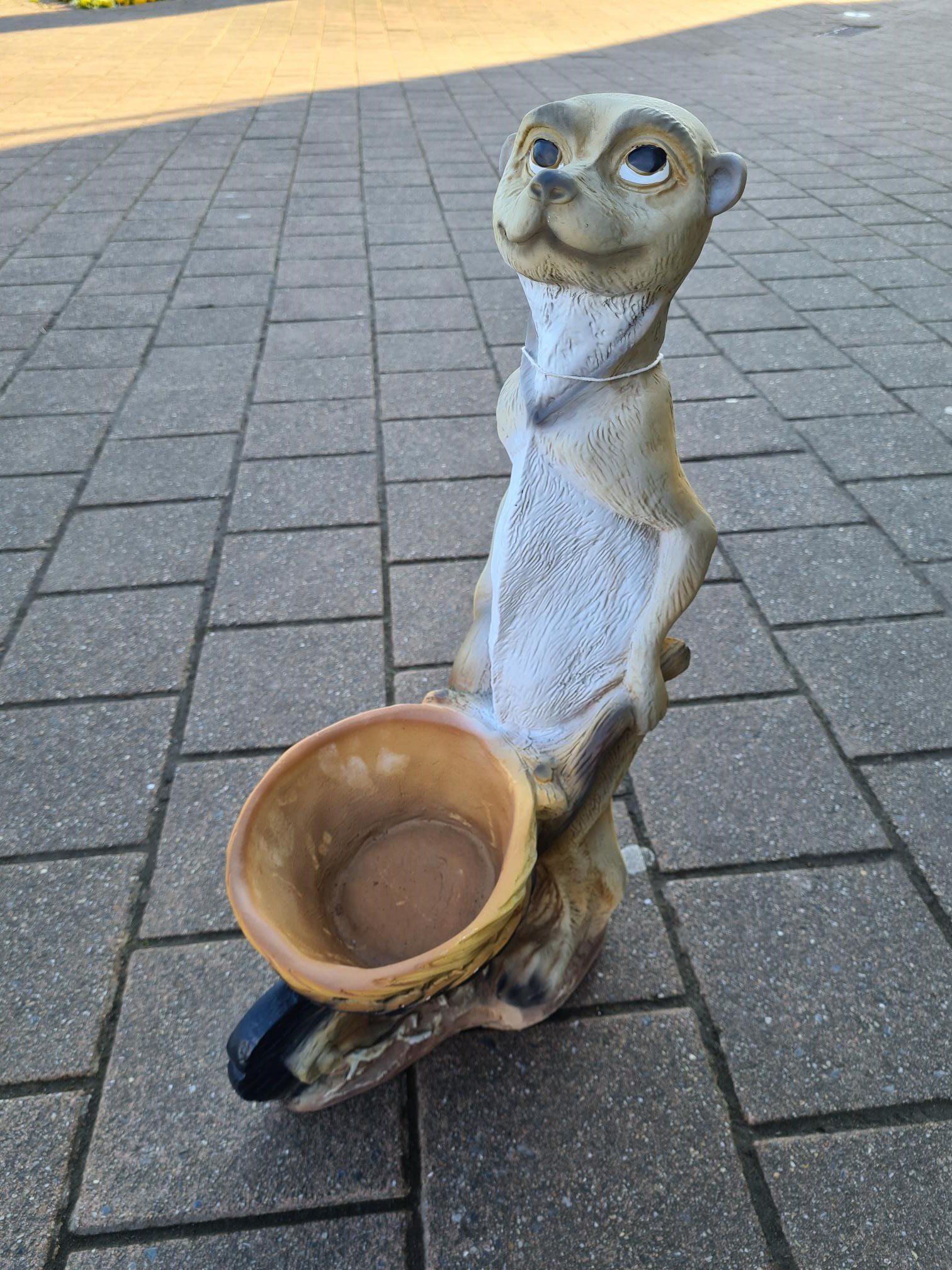 Klein stokstaartje-beeldje met een kom in zijn hand, staand op een geplaveide stoep buiten.