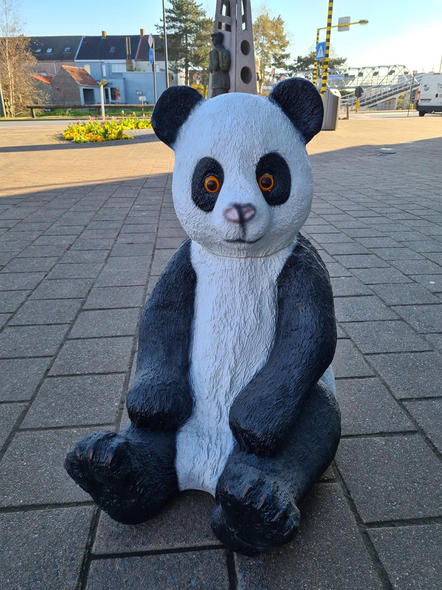 Een knuffelpanda zit op een geplaveide stoep buiten, voor gebouwen en bomen.