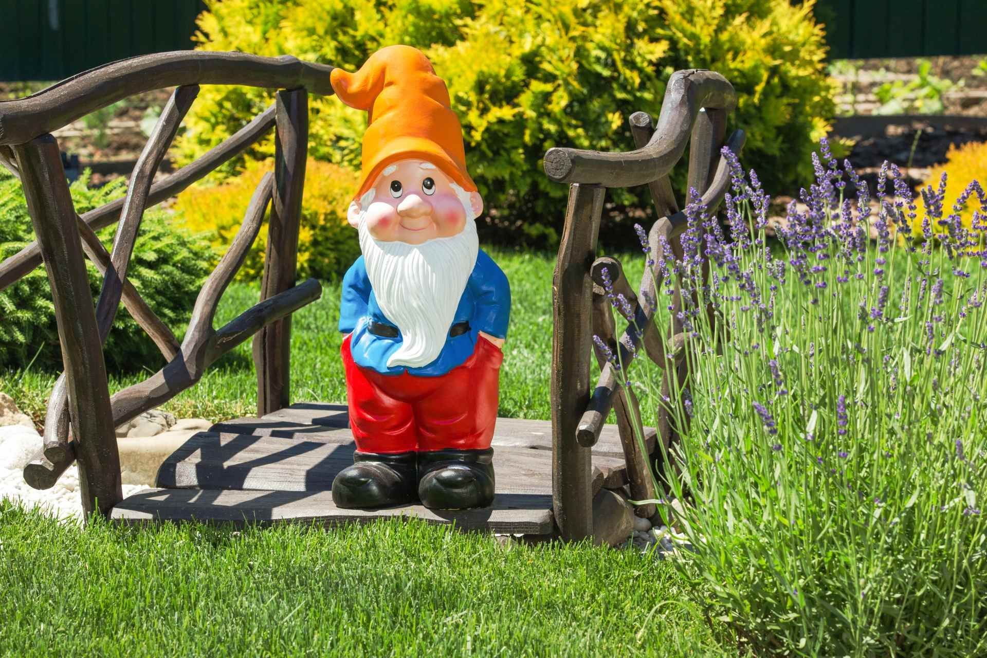 Een tuinkabouter met een oranje hoed staat op een klein houten bruggetje naast paarse bloemen.