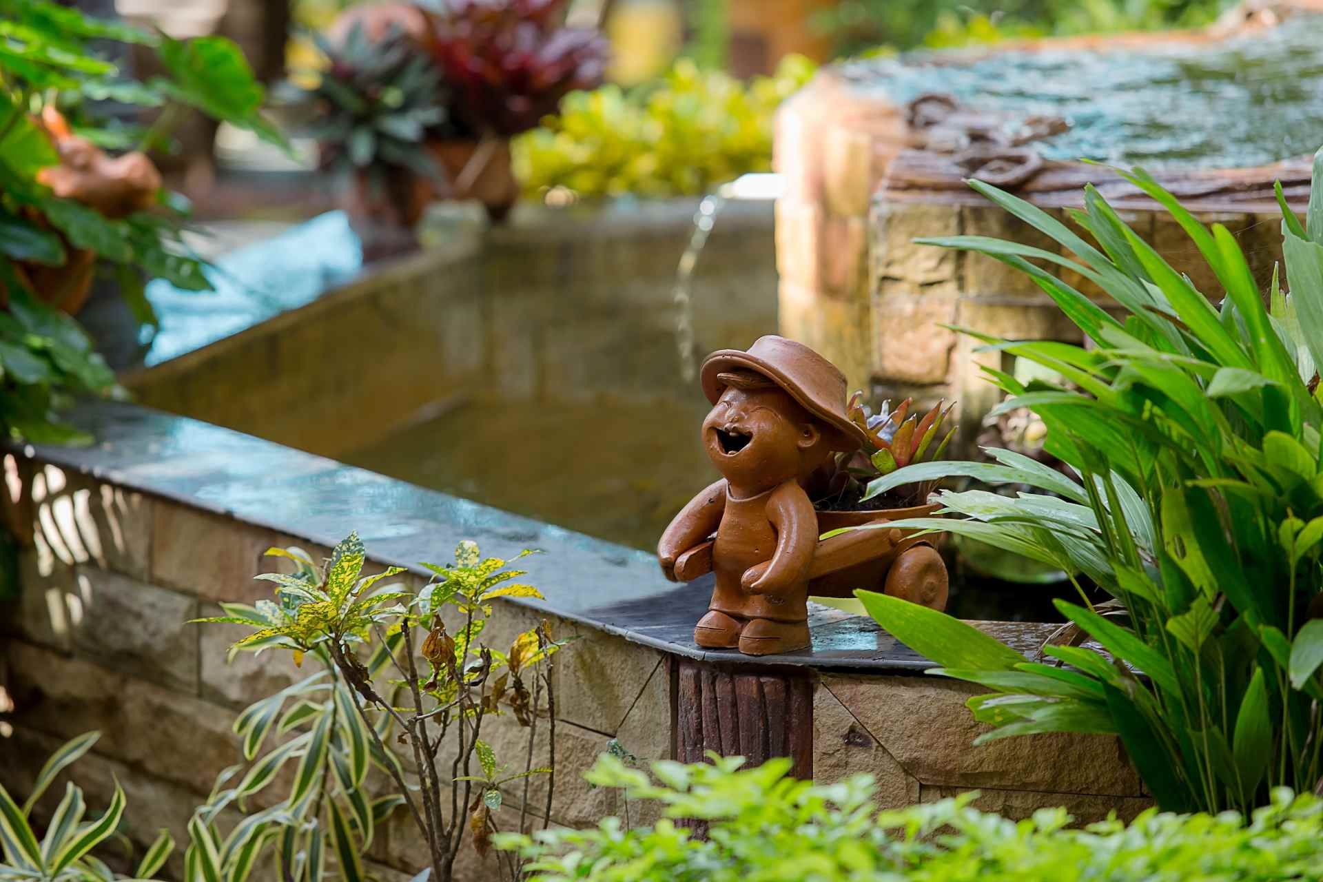 Een kleine tuinvijver met een piepklein bruin beeldje ernaast, omringd door weelderige groene planten en bloemen.
