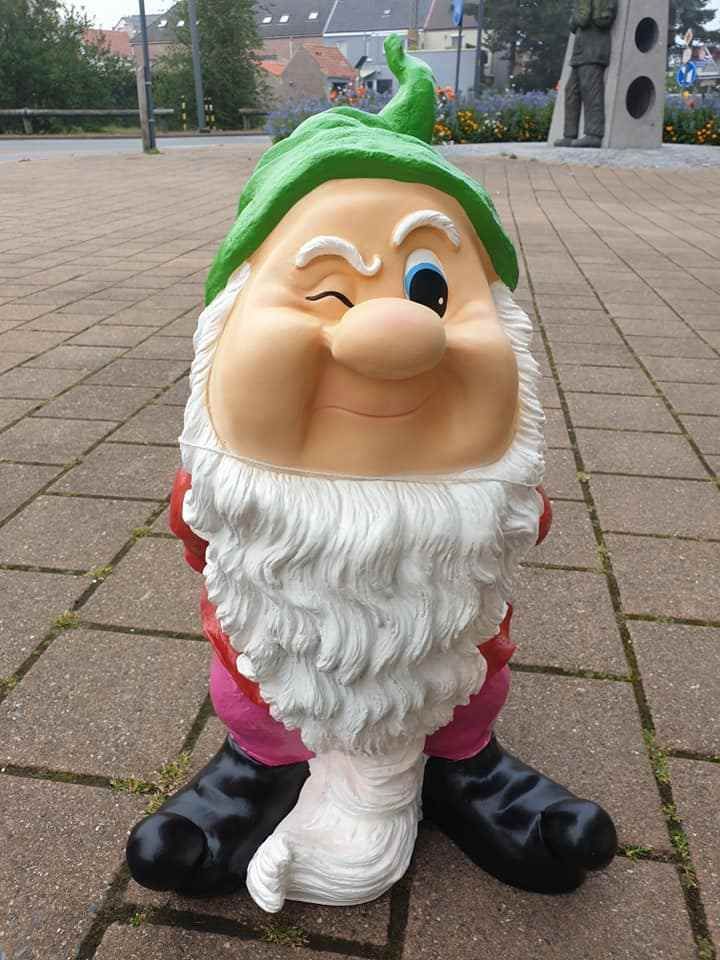 Een knipoogende tuinkabouter met groene hoed, witte baard, roze broek en zwarte laarzen op een bakstenen pad.