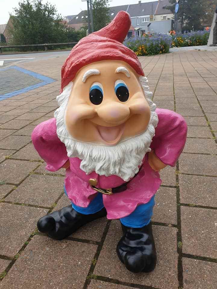 Een tuinkabouterbeeldje in een roze shirt, blauwe broek en rode hoed staat op een geplaveid pad.