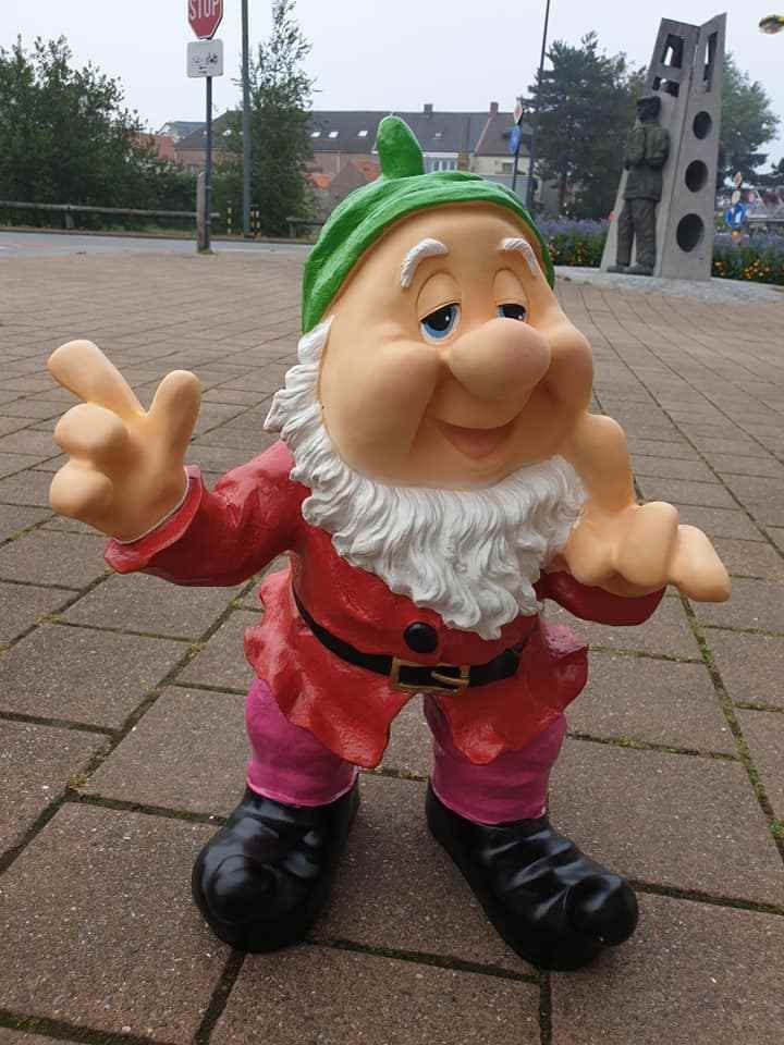 Een klein tuinkabouterbeeldje met groene hoed, rood shirt en zwarte laarzen staat op een geplaveid trottoir buiten.