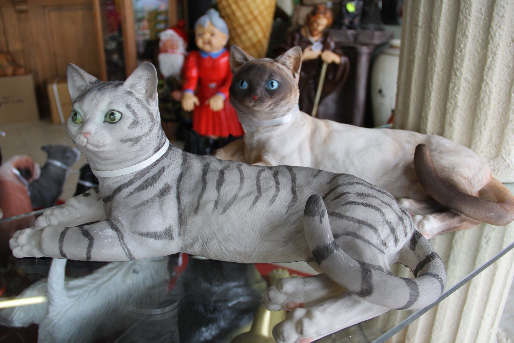 Twee grijze, gestreepte kattenbeeldjes liggen languit op een glazen plank met diverse decoratieve voorwerpen erachter.