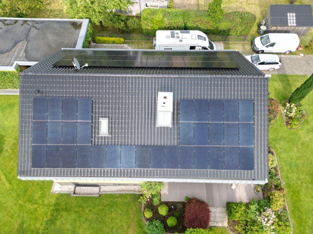 Draufsicht auf ein Haus mit Solarmodulen auf dem dunkelgrauen Dach, umgeben von grünem Rasen und einem kleinen Garten.