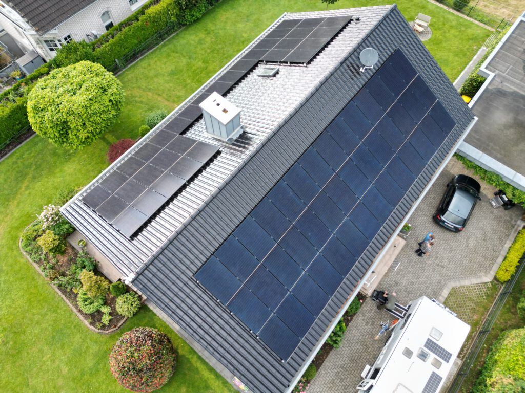 Solarmodule auf einem Wohnhausdach, umgeben von grünem Rasen, Bäumen und einem geparkten Auto.