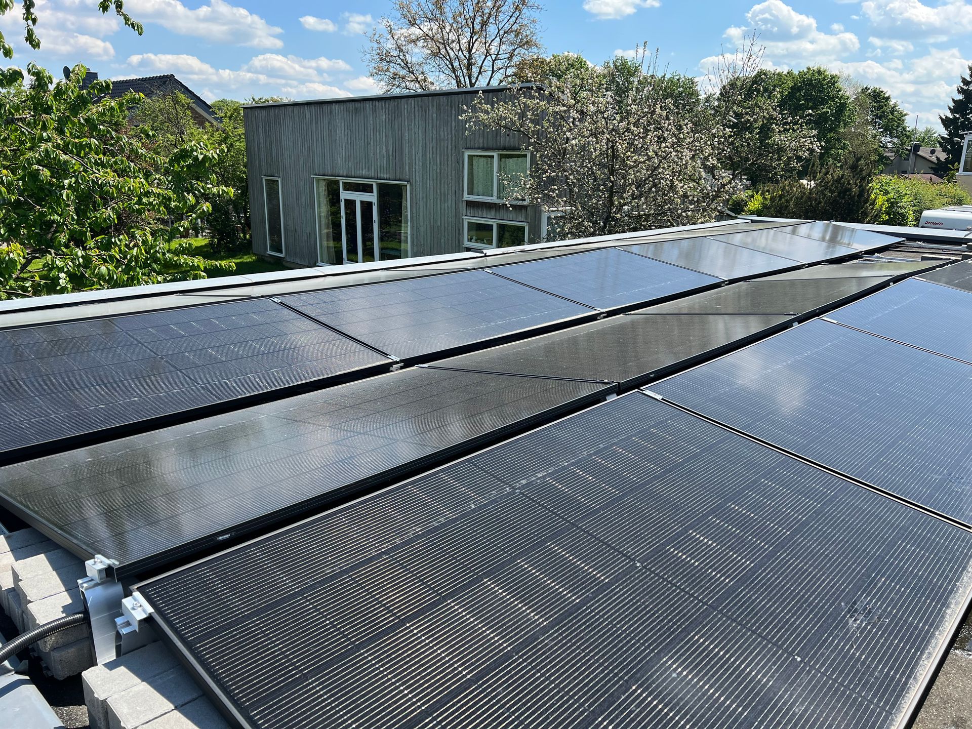 Solarpaneele auf dem Dach eines kleinen Gebäudes, im Hintergrund Bäume und blauer Himmel.