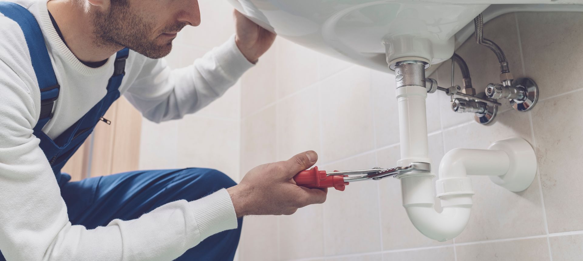 Un plombier utilise une clé pour travailler sur les tuyaux sous un lavabo blanc dans une salle de bains.