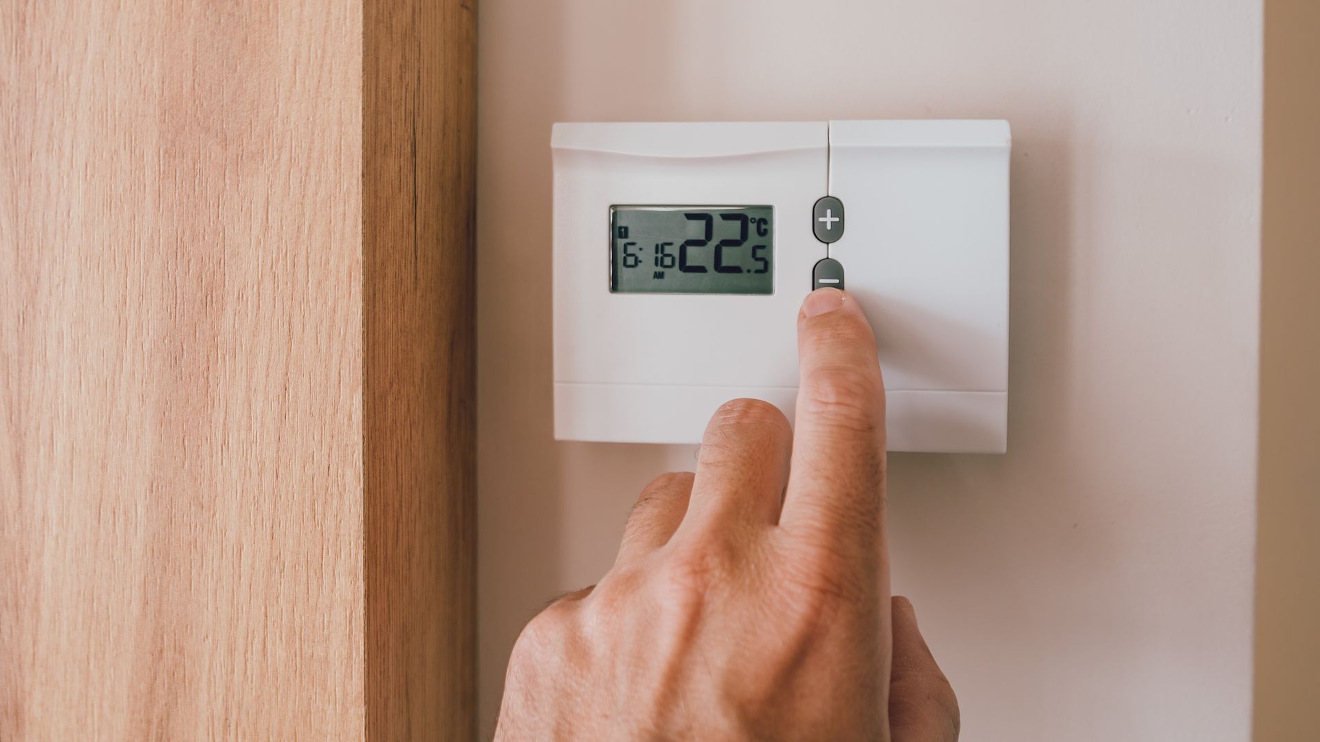 Une main ajuste un thermostat sur un mur blanc et le règle sur 22,5 degrés. Lambris à gauche.