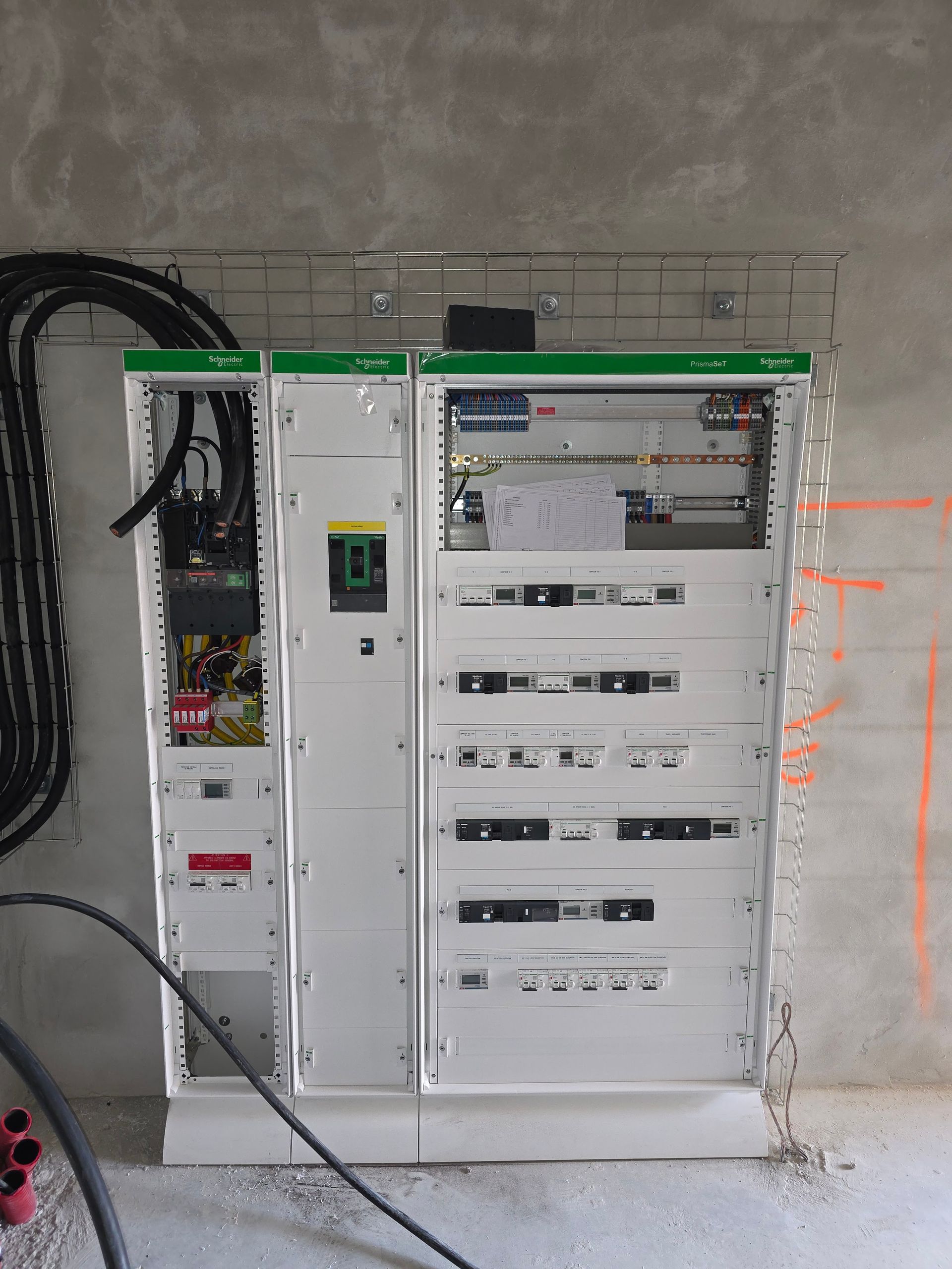 Un électricien en salopette et casque de chantier inspecte le câblage d'une armoire électrique à l'aide d'une tablette.