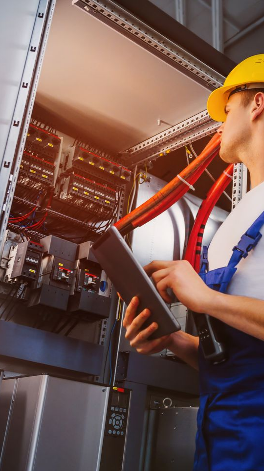 Un électricien en salopette et casque de chantier inspecte le câblage d'une armoire électrique à l'aide d'une tablette.