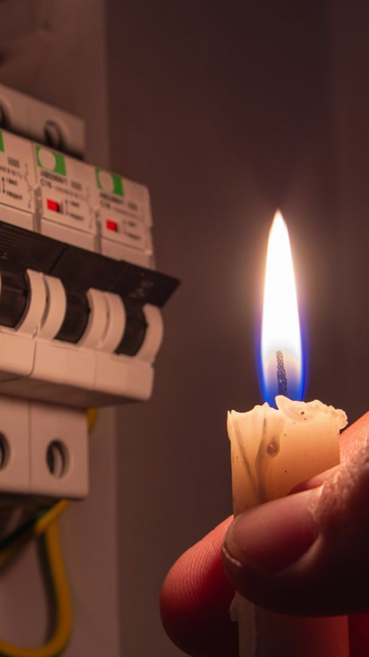 Une personne tient une bougie allumée près d'un panneau électrique ; lumières éteintes.