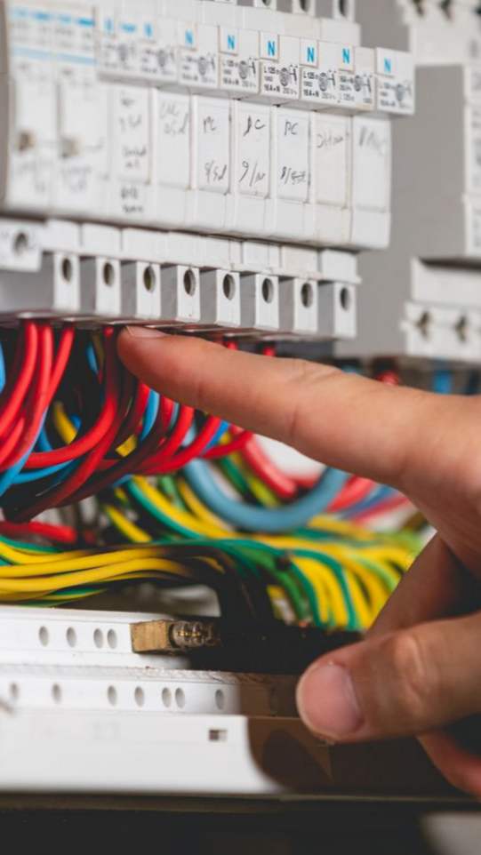 Main pointant du doigt des fils électriques colorés reliés à un tableau de disjoncteurs.