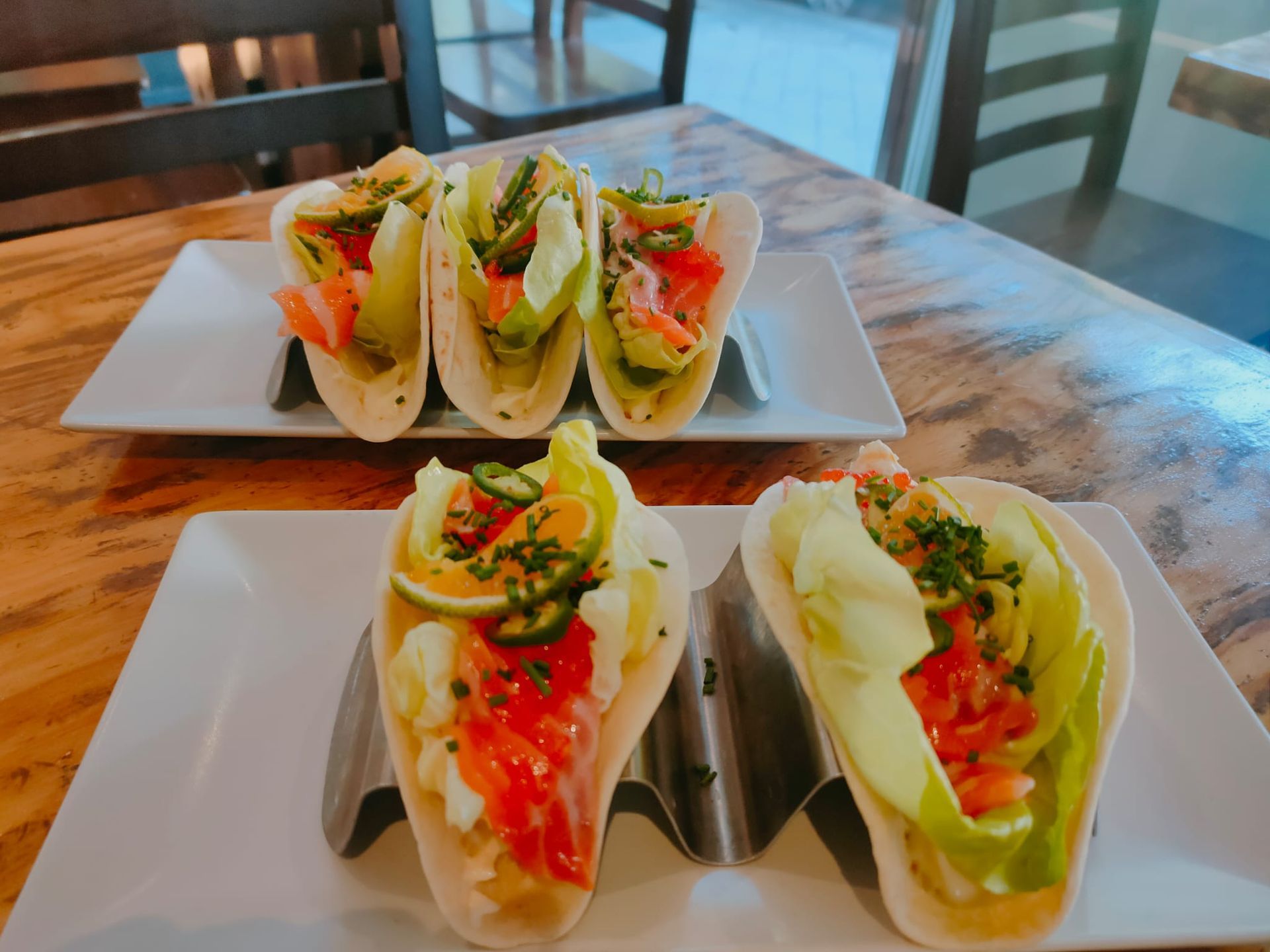 Tacos en platos blancos con rellenos coloridos sobre una mesa de madera en un comedor luminoso.