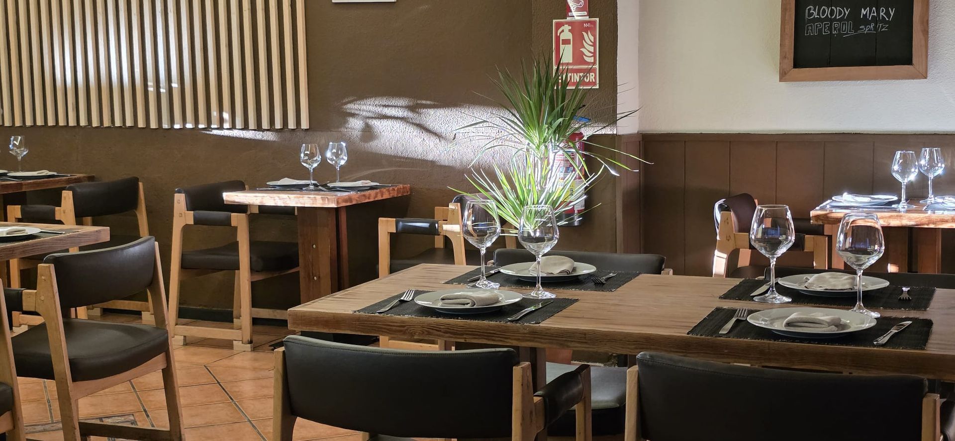 El interior de un restaurante con varias mesas de madera dispuestas con copas de vino, platos y sillas negras.