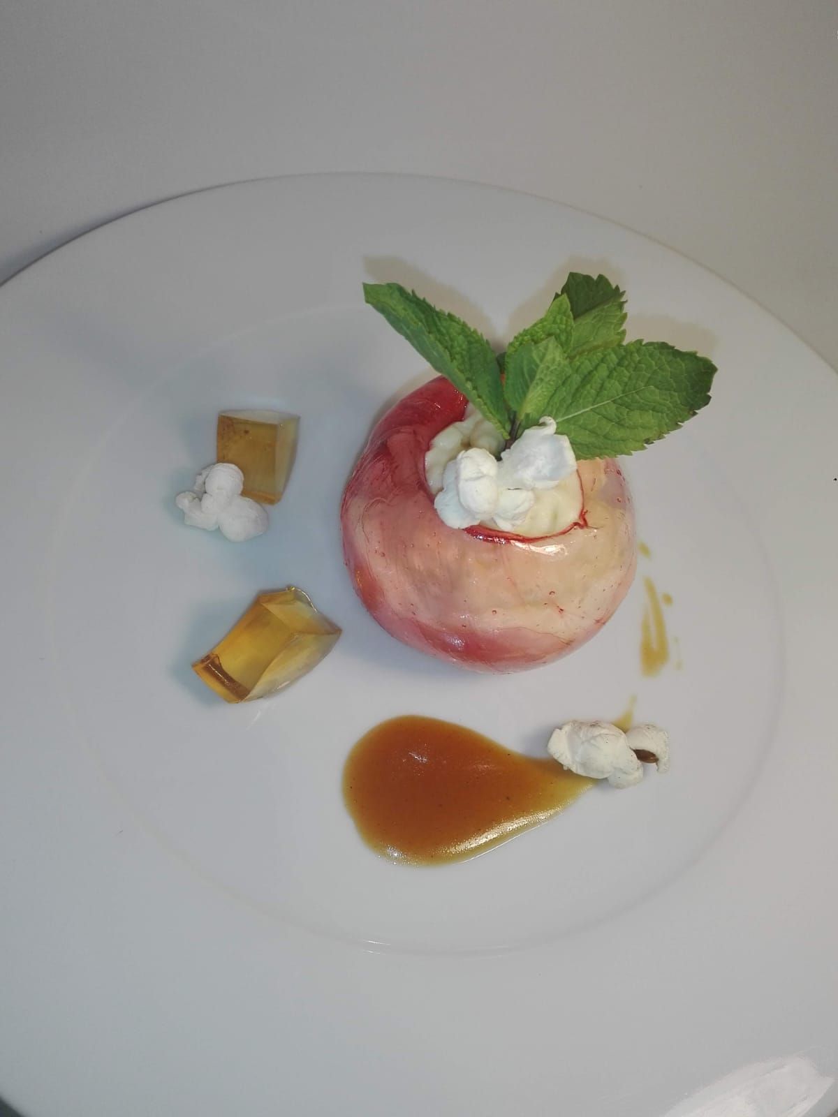 Un postre de manzana al horno adornado con menta, crema batida, pequeños cubos de fruta y un chorrito de salsa de caramelo, servido en un plato blanco.