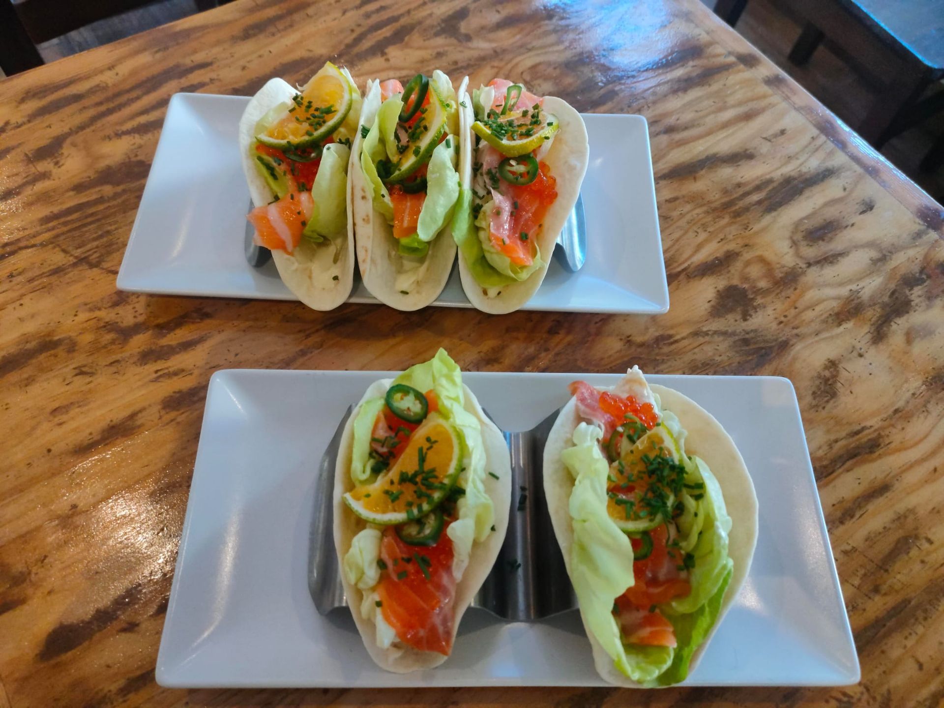 Cinco tacos de salmón adornados con verduras y rodajas de jalapeño, dispuestos en dos bandejas blancas sobre una mesa de madera.