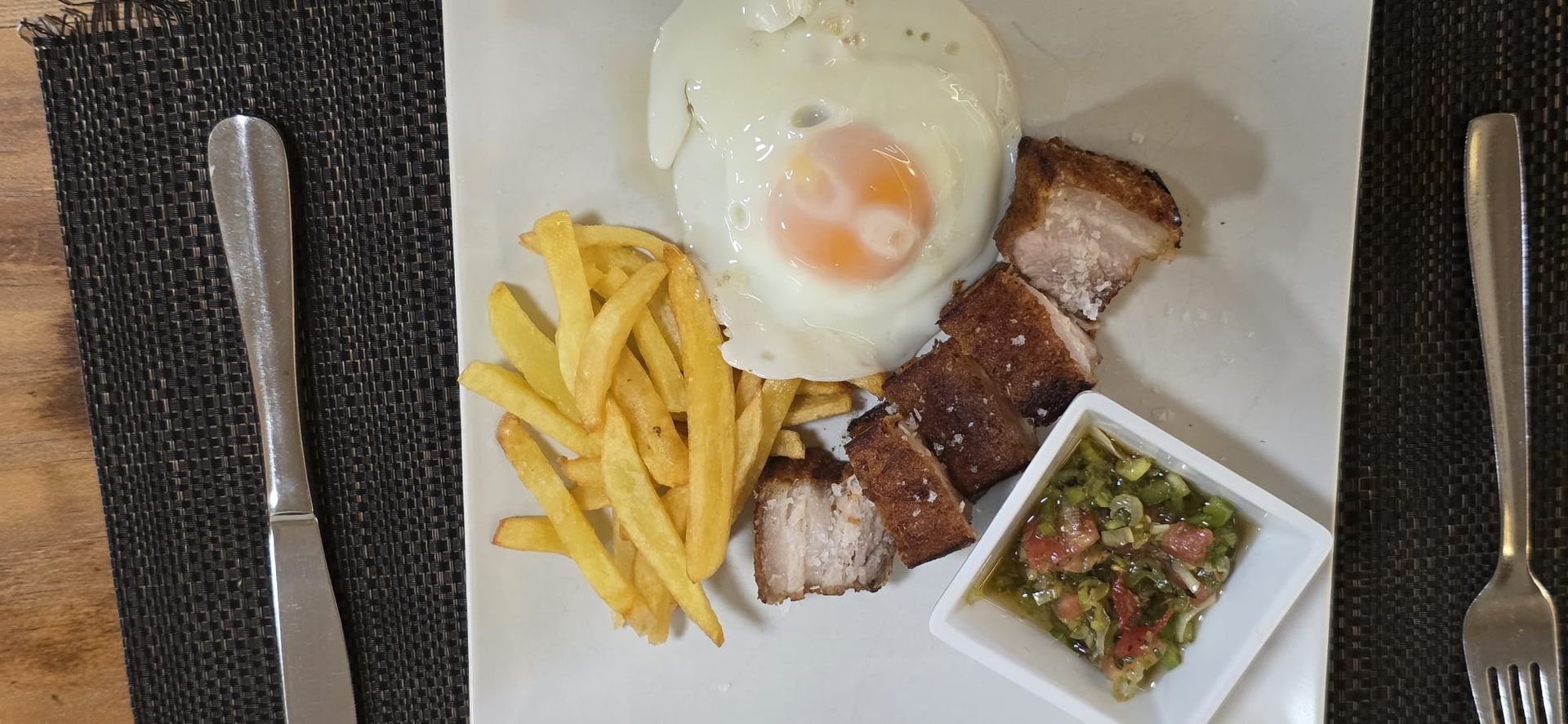 Un plato con huevo frito, patatas fritas, panceta de cerdo en lonchas y una guarnición de salsa, servido con tenedor y cuchillo sobre un mantel individual.