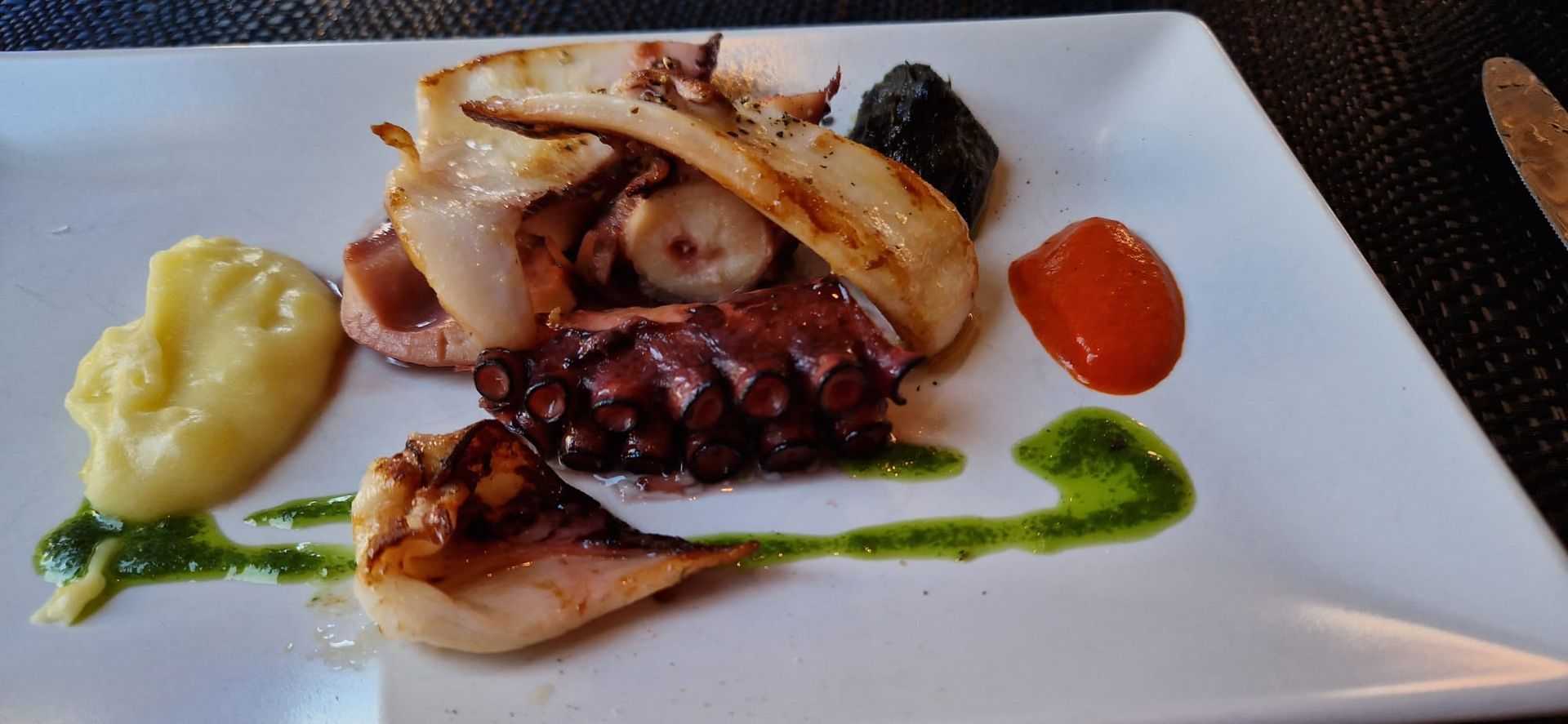 Pulpo a la parrilla servido en un plato blanco con puré de patatas, salsa de pimiento rojo y un chorrito de aceite de hierbas verdes.