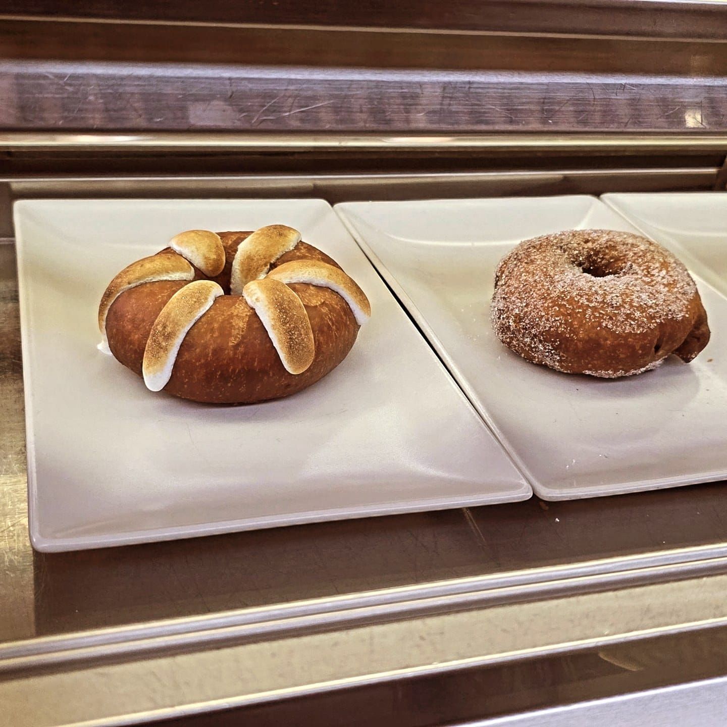 En una vitrina, dos rosquillas reposan sobre platos rectangulares blancos; una está cubierta con malvavisco tostado y la otra con azúcar.
