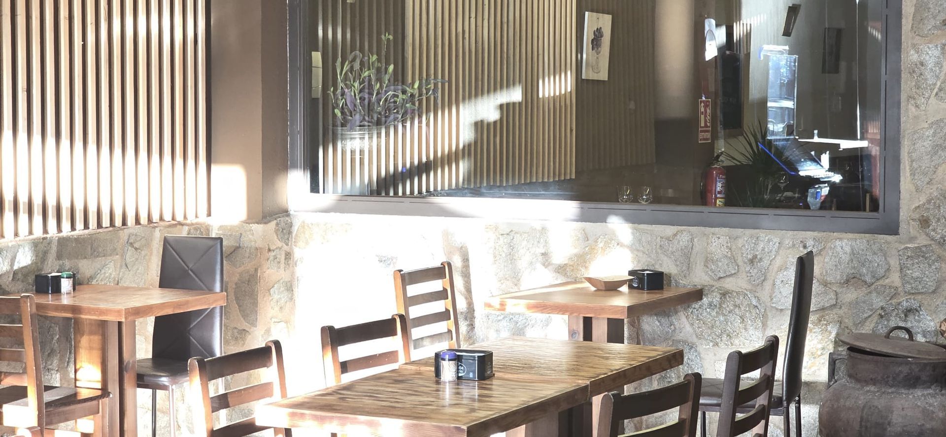 Un comedor rústico al aire libre con varias mesas y sillas de madera, situado contra una pared de piedra con persianas verticales.