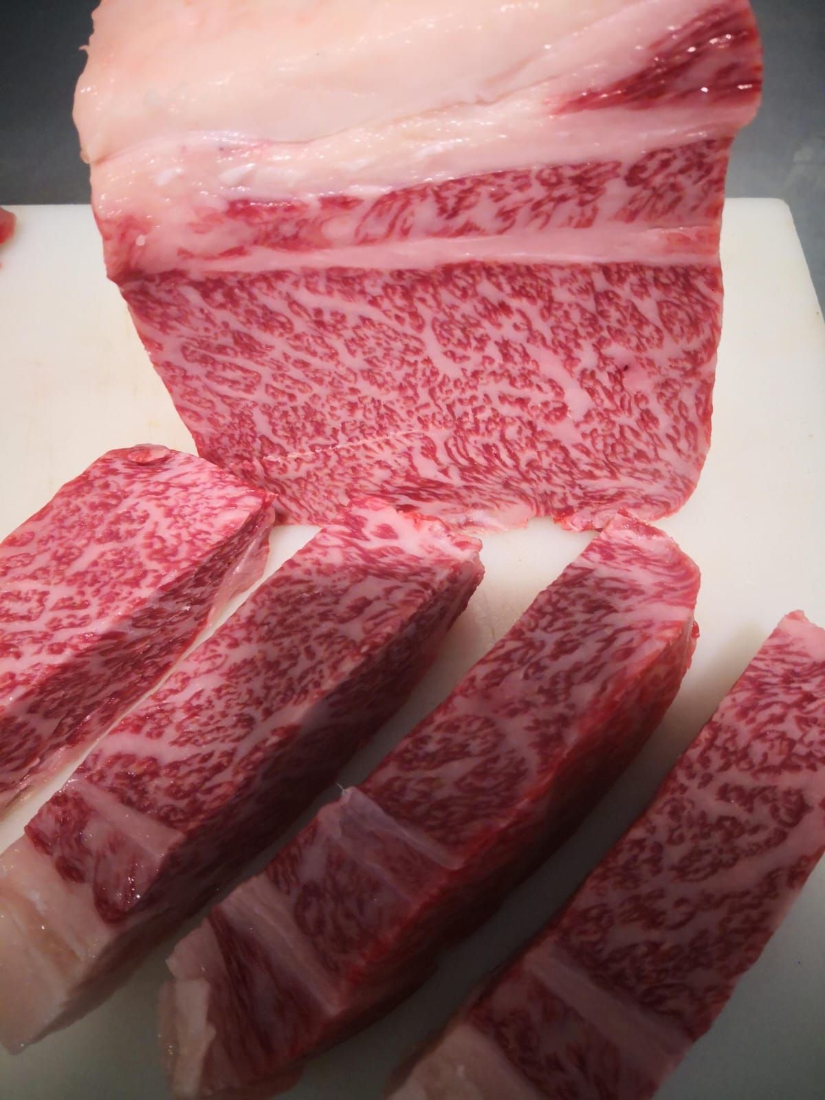 Un bloque de carne de Wagyu cruda, con alto grado de marmoleado, y cuatro rebanadas gruesas dispuestas sobre una tabla de cortar blanca.
