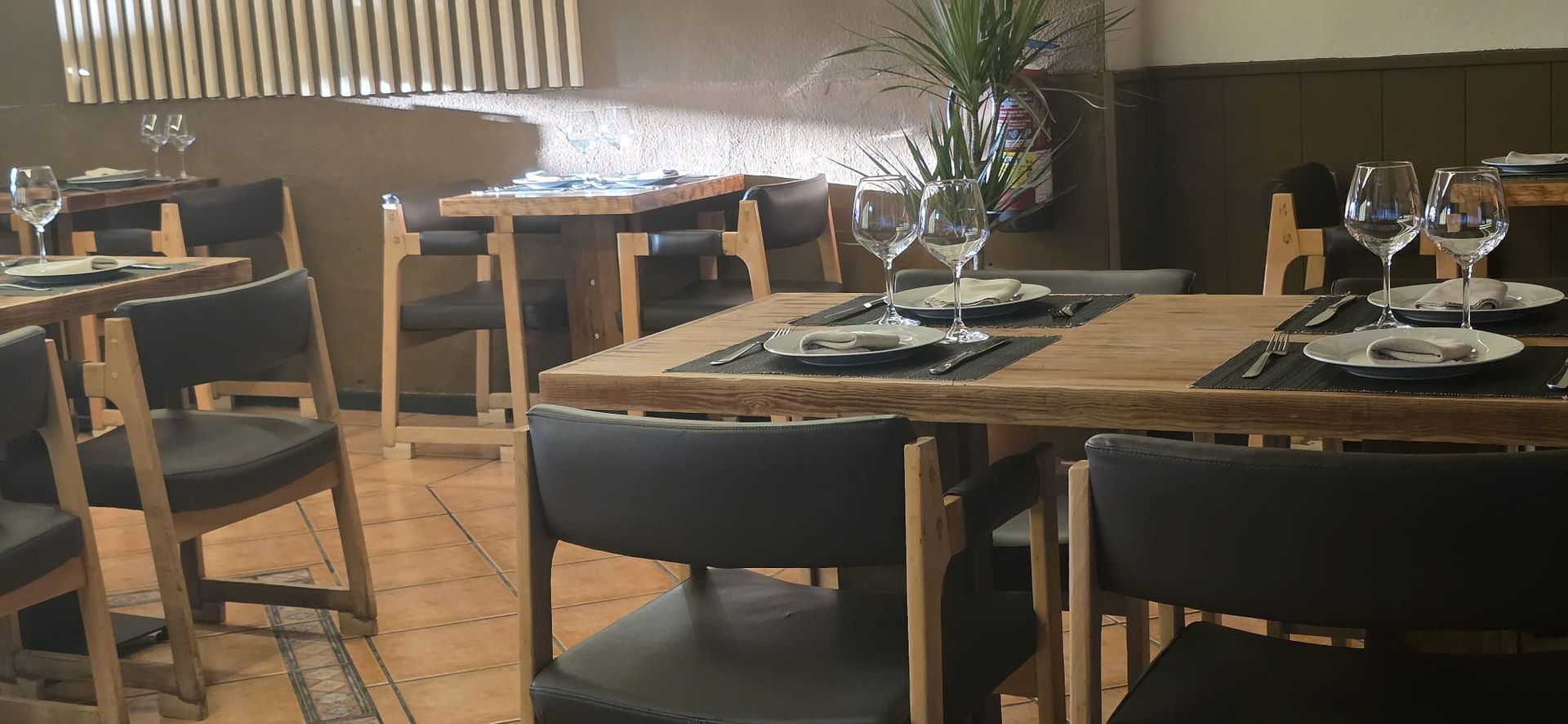 El interior de un restaurante cuenta con varias mesas de madera con vajilla, copas de vino y sillas tapizadas en tonos oscuros.