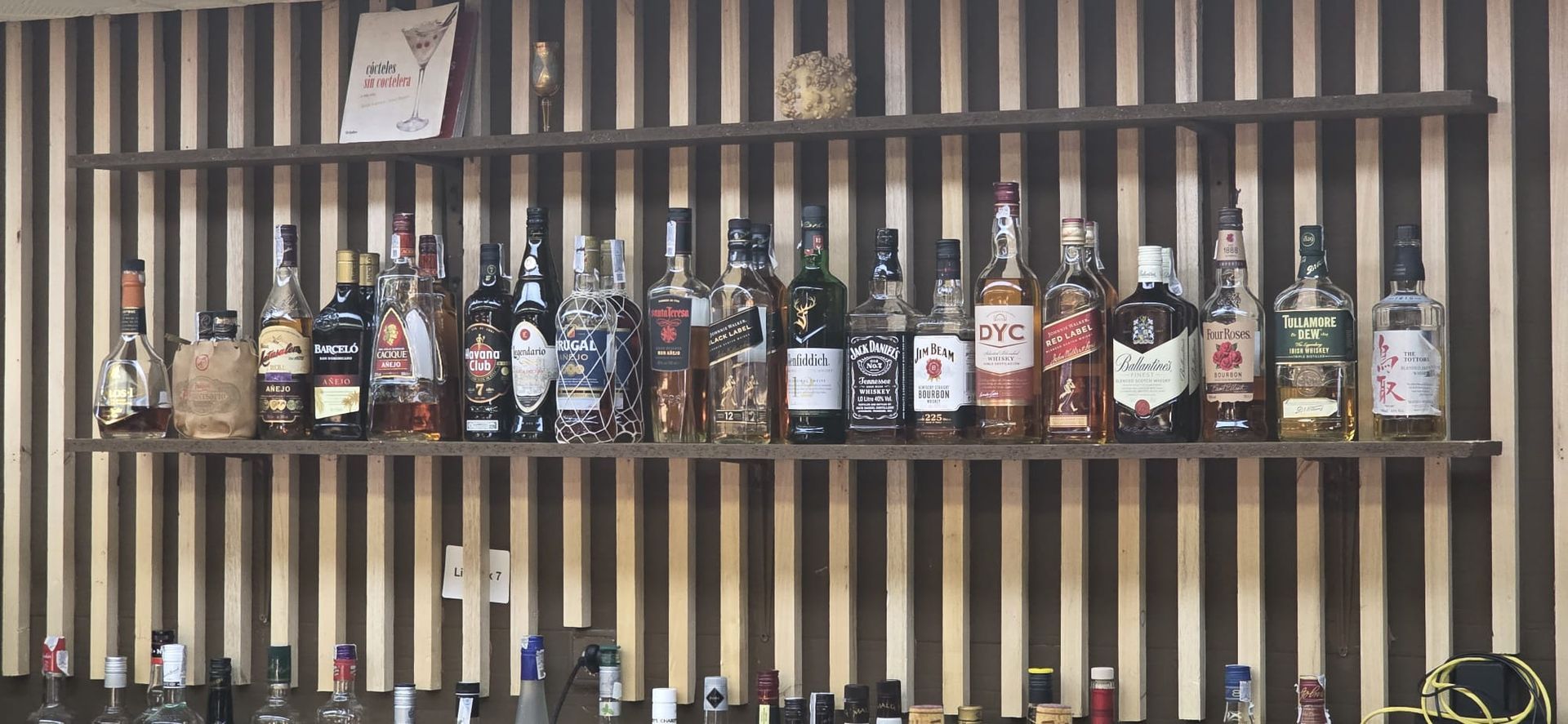 Una hilera de botellas de licor de diferentes tipos expuestas en un estante de madera contra una pared de listones verticales.