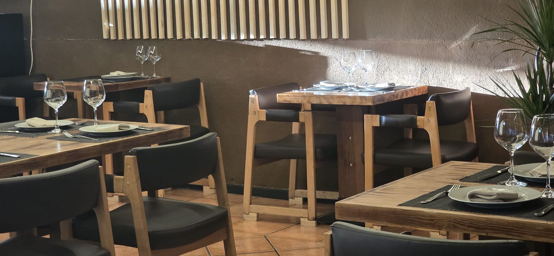 Mesas vacías dispuestas para comer en un restaurante con muebles de madera y sillas oscuras contra una pared texturizada.