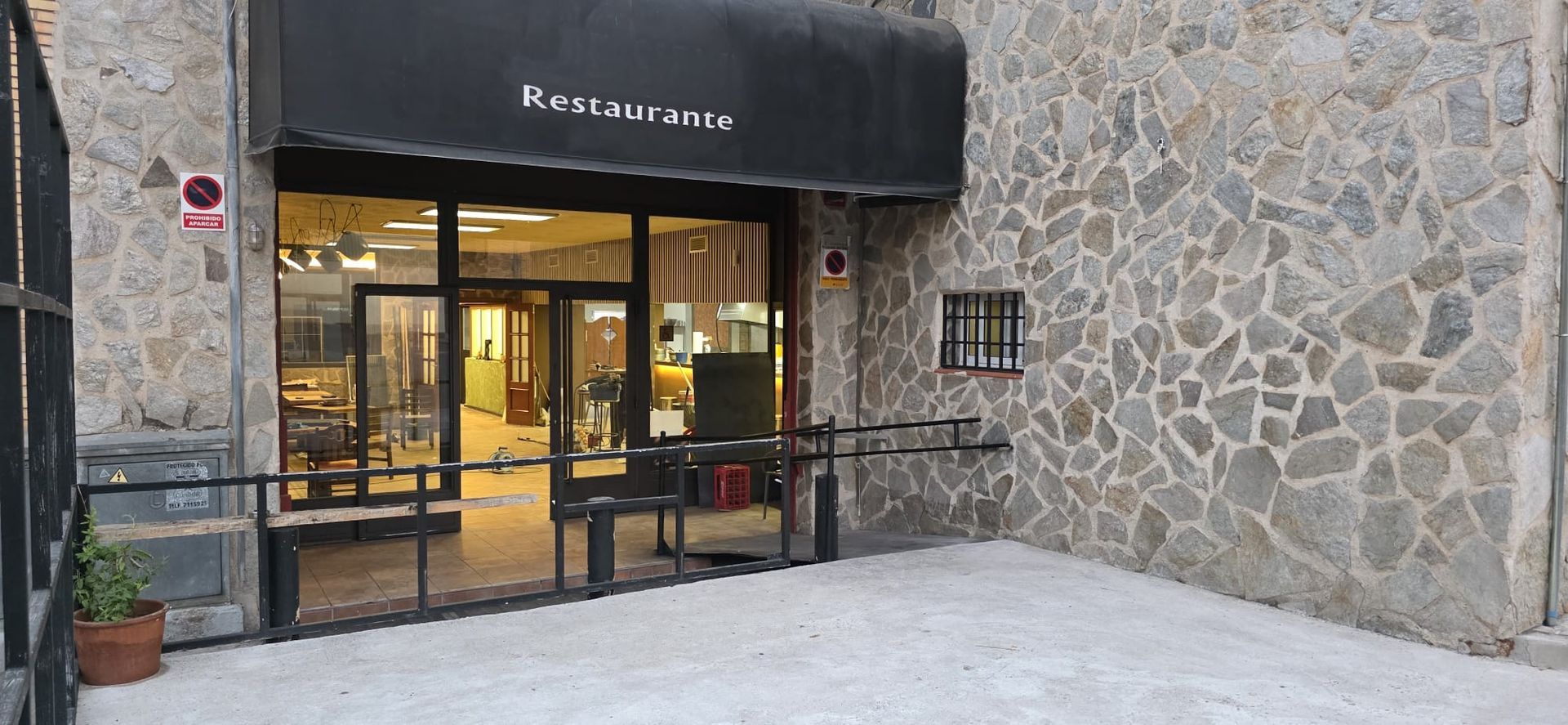 Entrada a un restaurante con toldo negro y fachada de piedra, que cuenta con puerta de cristal y barandillas de seguridad metálicas.