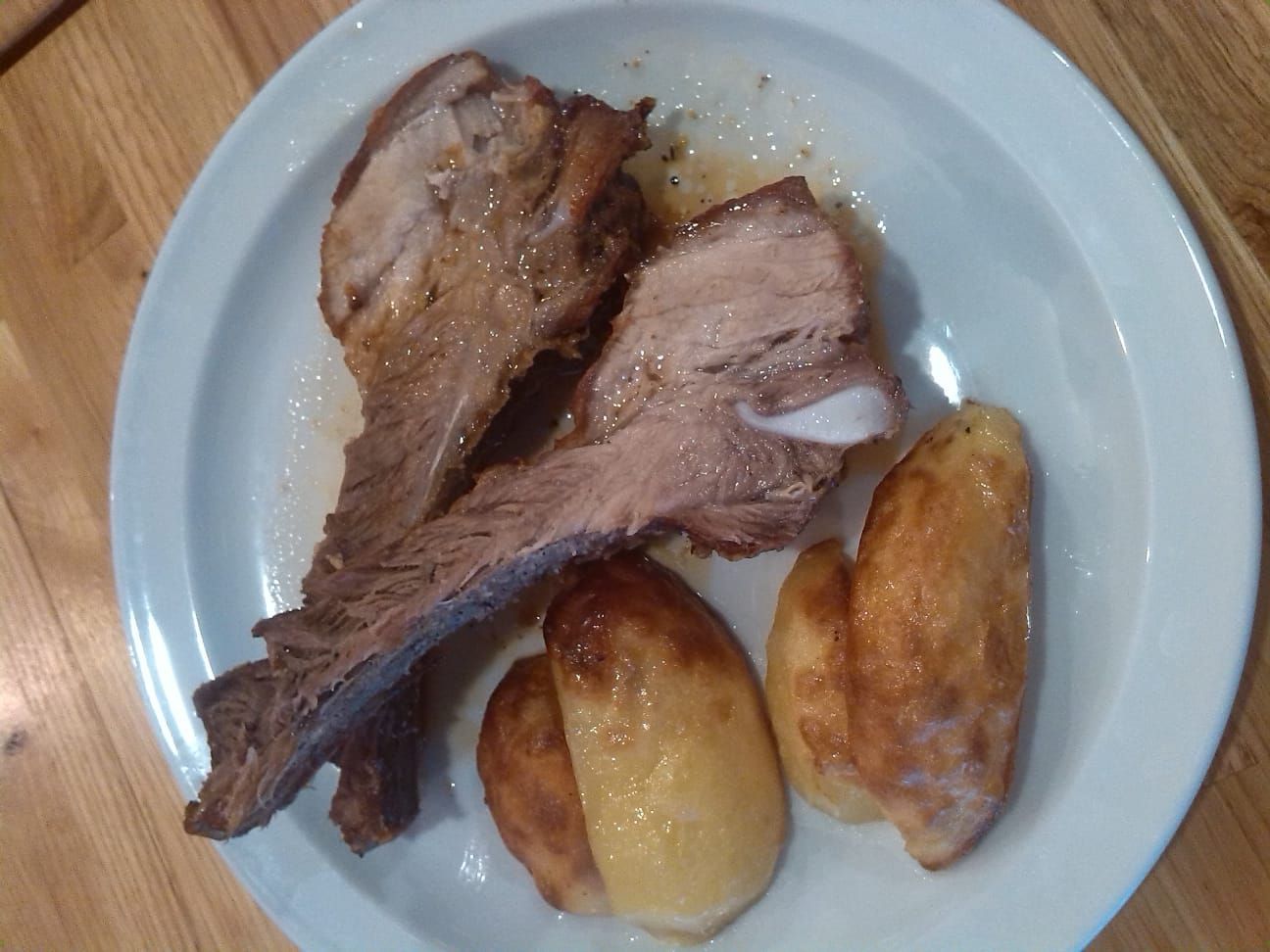 Un plato blanco con dos trozos de carne cocida, posiblemente costillas de cerdo, junto a tres patatas asadas de color marrón dorado.