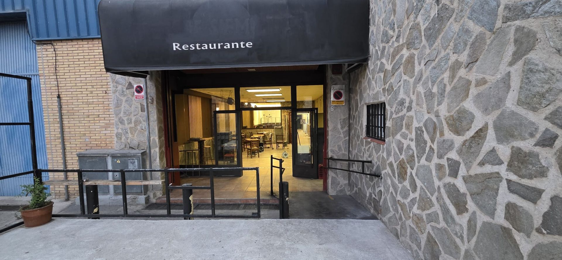 Entrada de restaurante con toldo negro y fachada de piedra, con puerta de cristal y barandilla metálica exterior.