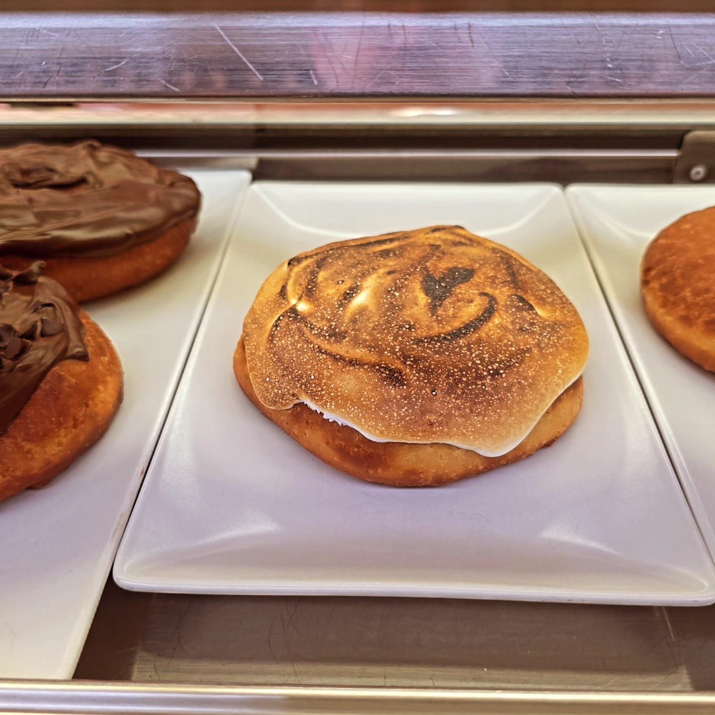 Primer plano de un pastel estilo s'mores con cobertura de malvavisco tostado sobre un plato blanco en una vitrina de una panadería.