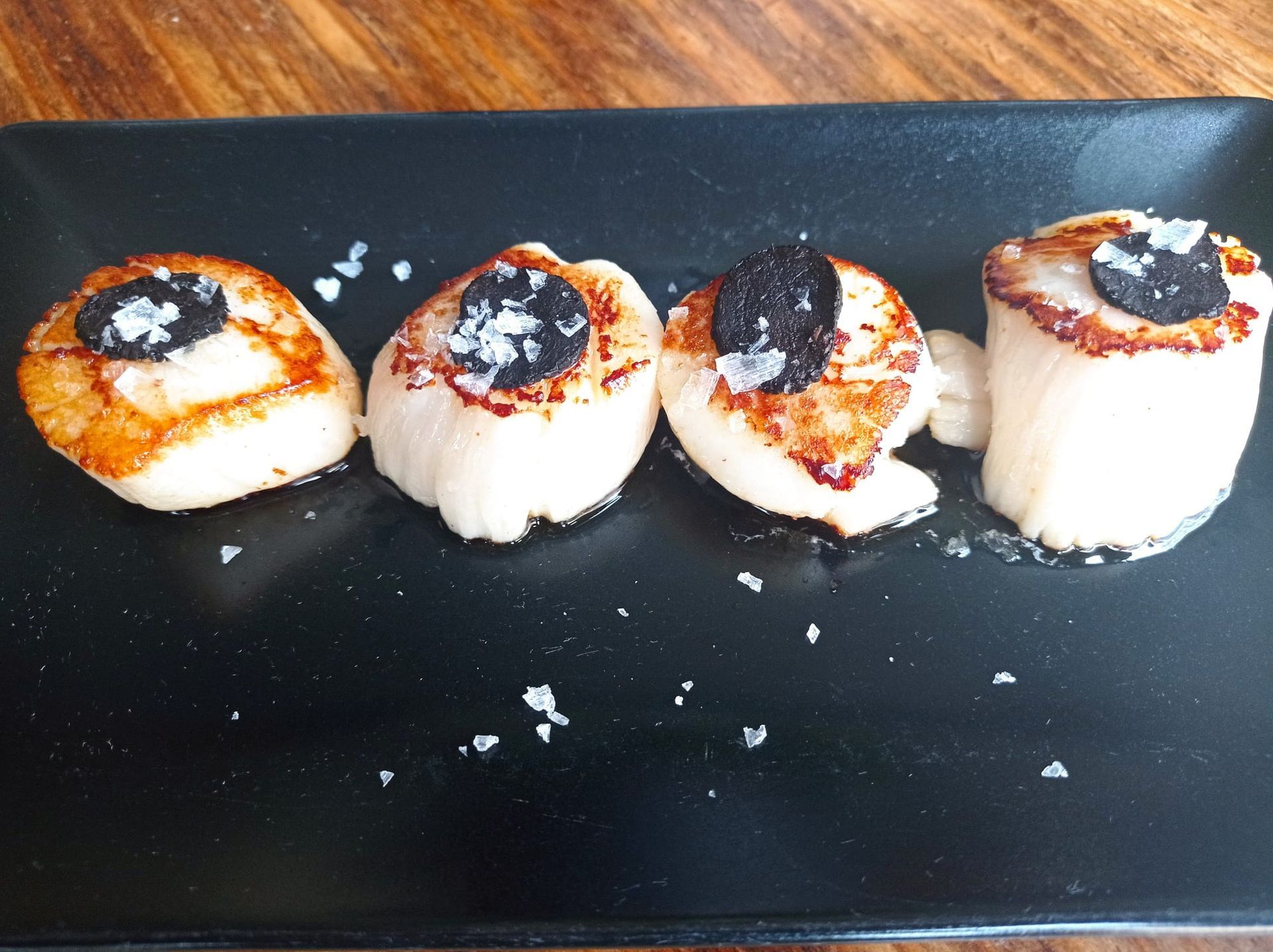 Cuatro vieiras selladas a la plancha, cubiertas con láminas de trufa negra y sal marina gruesa, servidas en un plato rectangular negro.