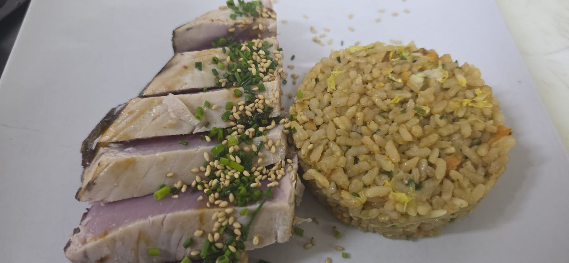 Filete de atún sellado en rodajas, cubierto con semillas de sésamo y hierbas, servido con una porción de arroz frito en un plato blanco.