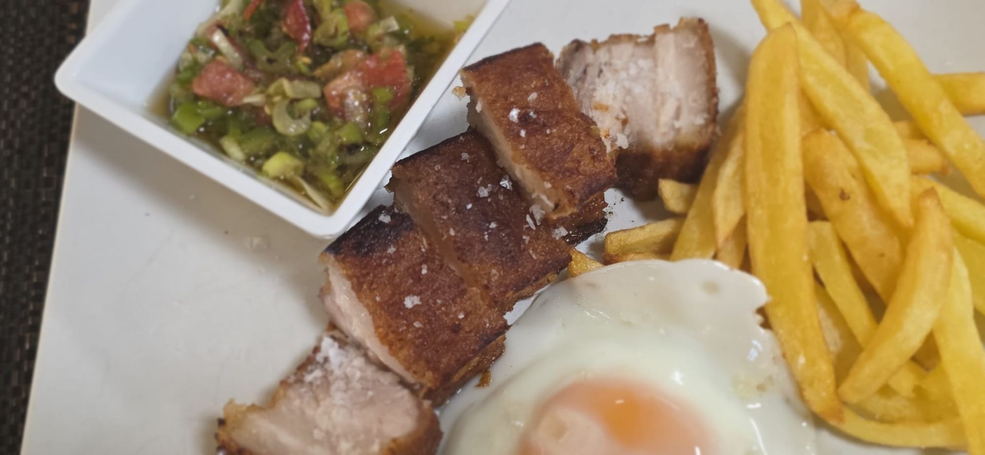 Un plato con panceta de cerdo crujiente, un huevo frito, patatas fritas doradas y un pequeño recipiente de salsa verde.