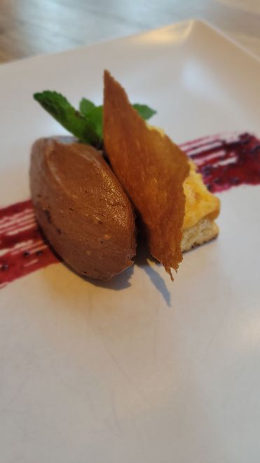 Un postre emplatado con una quenelle de mousse de chocolate, una oblea crujiente, un pequeño bizcocho, menta y un remolino de coulis de frutos rojos.